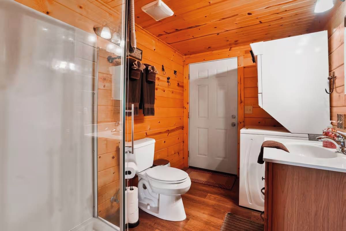 Baño de cabaña de madera con mampara de ducha de vidrio, inodoro, lavamanos y puerta blanca.