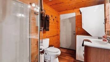 Baño de cabaña de madera con mampara de ducha de vidrio, inodoro, lavamanos y puerta blanca.