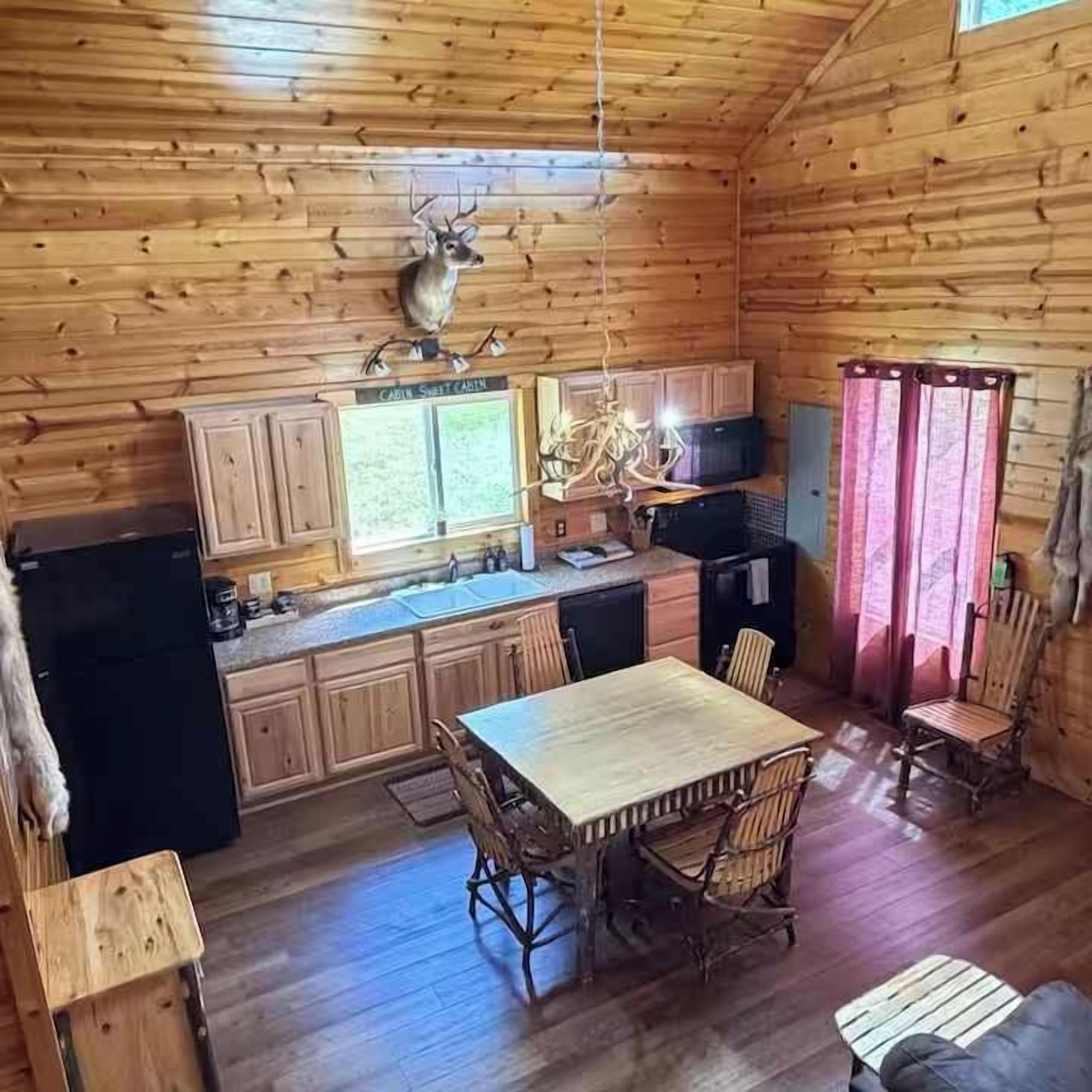 A wooden cabin interior with a dining table, chairs, a refrigerator, and a mounted deer head.