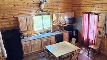 A wooden cabin interior with a dining table, chairs, a refrigerator, and a mounted deer head.