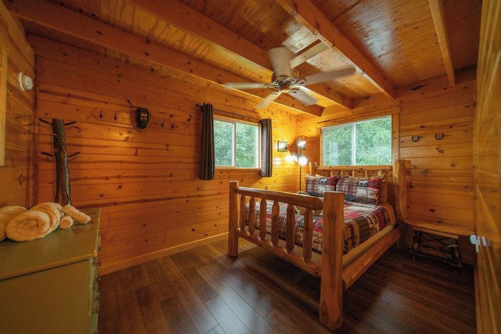 A wooden cabin room with a bed, a ceiling fan, and two windows.