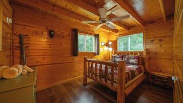 A wooden cabin room with a bed, a ceiling fan, and two windows.