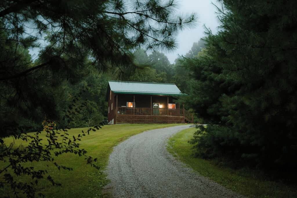 A cabin with a porch, surrounded by trees and a gravel path leading to it.