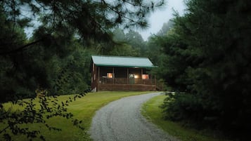 A cabin with a porch, surrounded by trees and a gravel path leading to it.