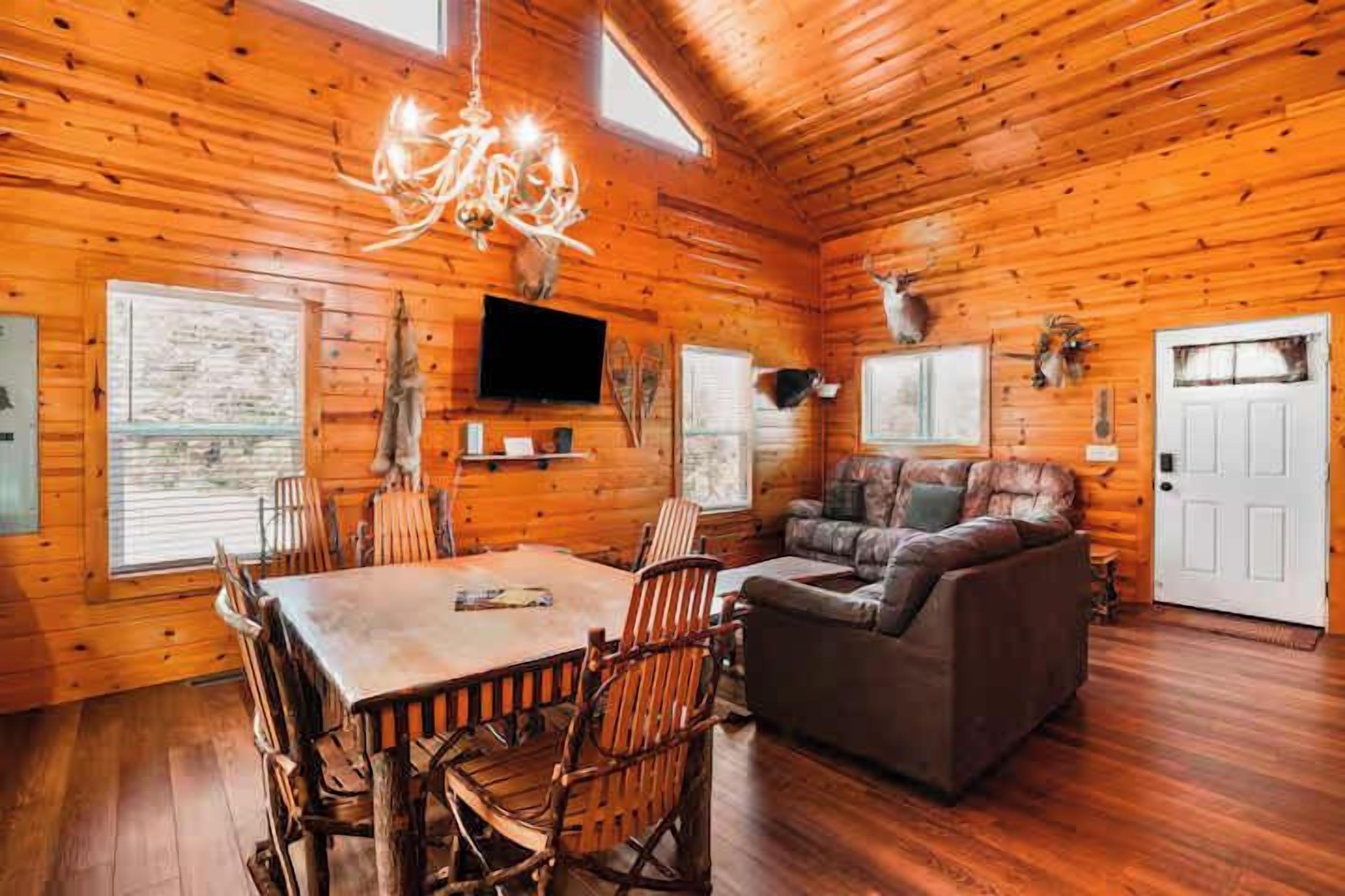 A log cabin interior with a dining table, chairs, a sofa, a chandelier, mounted television, and deer antlers on the walls.