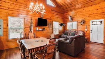 A log cabin interior with a dining table, chairs, a sofa, a chandelier, mounted television, and deer antlers on the walls.