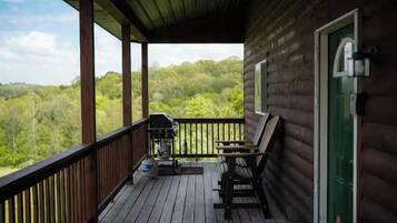 Una cabaña de madera con un porche, una parrilla y vistas al bosque.