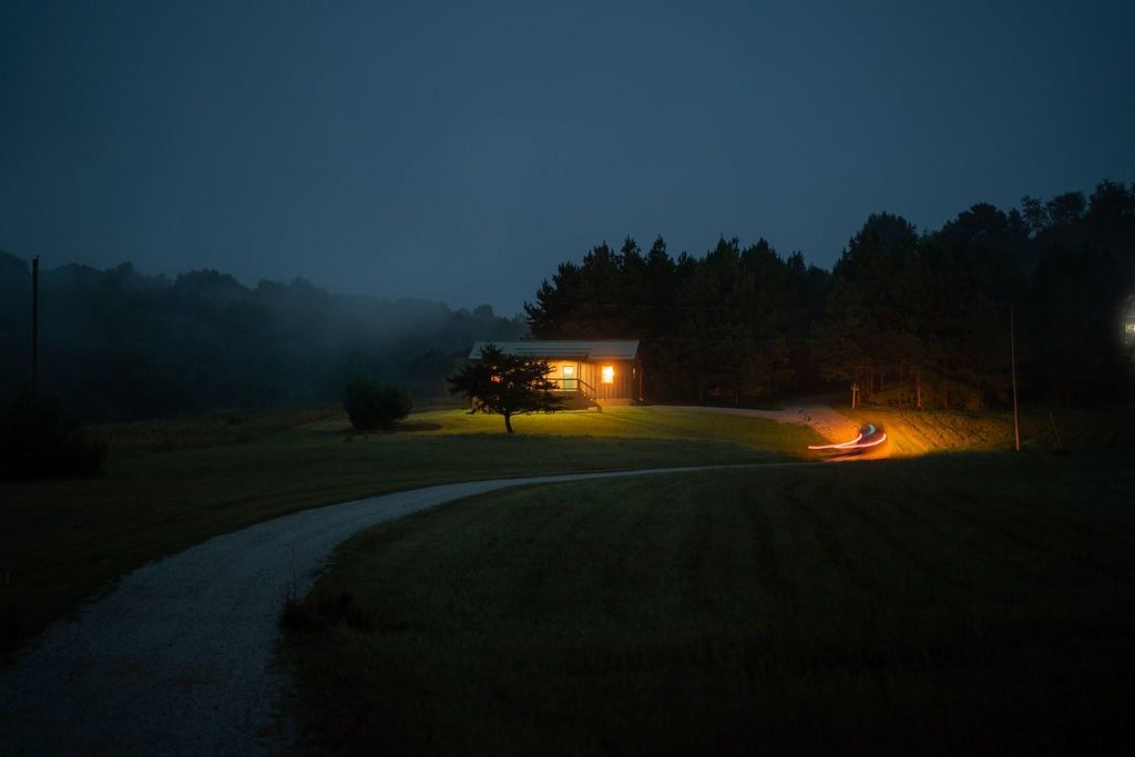A house with a porch and a driveway leading to it, surrounded by trees and a grassy area.