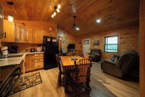 Interior de una cabaña con una mesa de comedor de madera, sillas, una cocina, un sofá y una ventana.