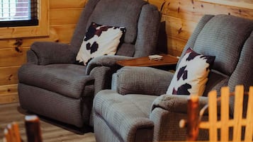A cozy room with two grey upholstered chairs with cowhide pillows, a small wooden table, and a window with blinds.