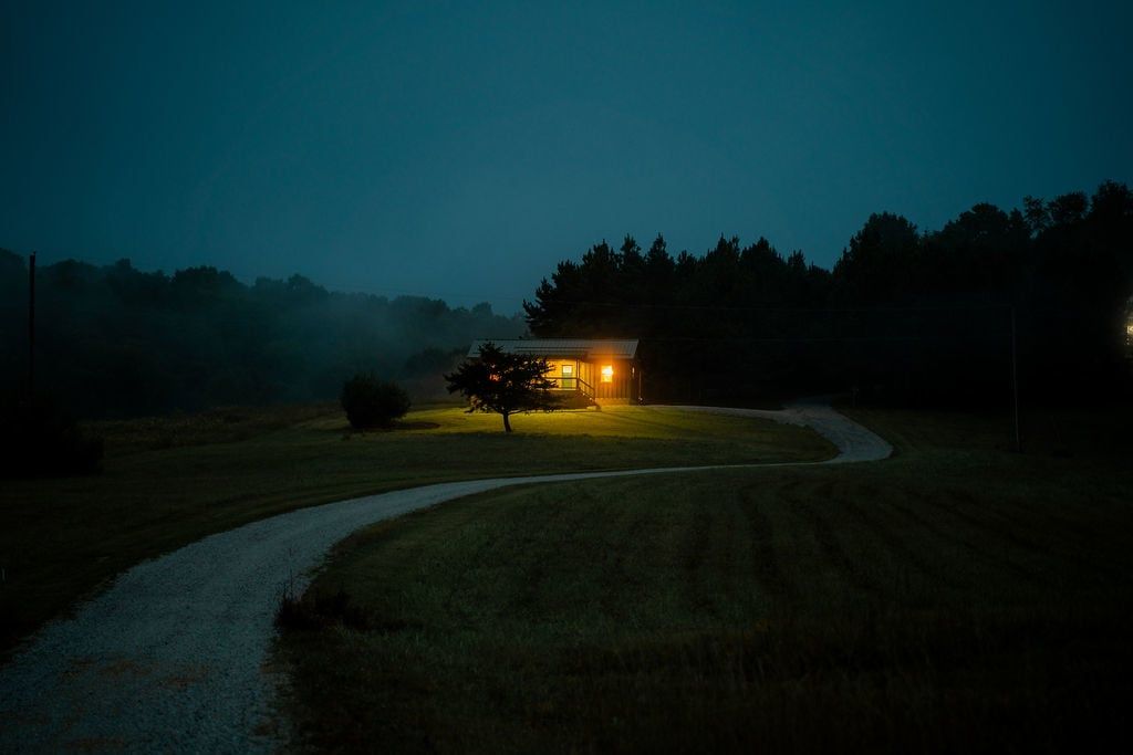 A rural landscape at dusk with a house illuminated by warm light, surrounded by trees and a winding path.