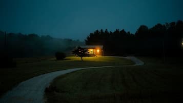 A rural landscape at dusk with a house illuminated by warm light, surrounded by trees and a winding path.