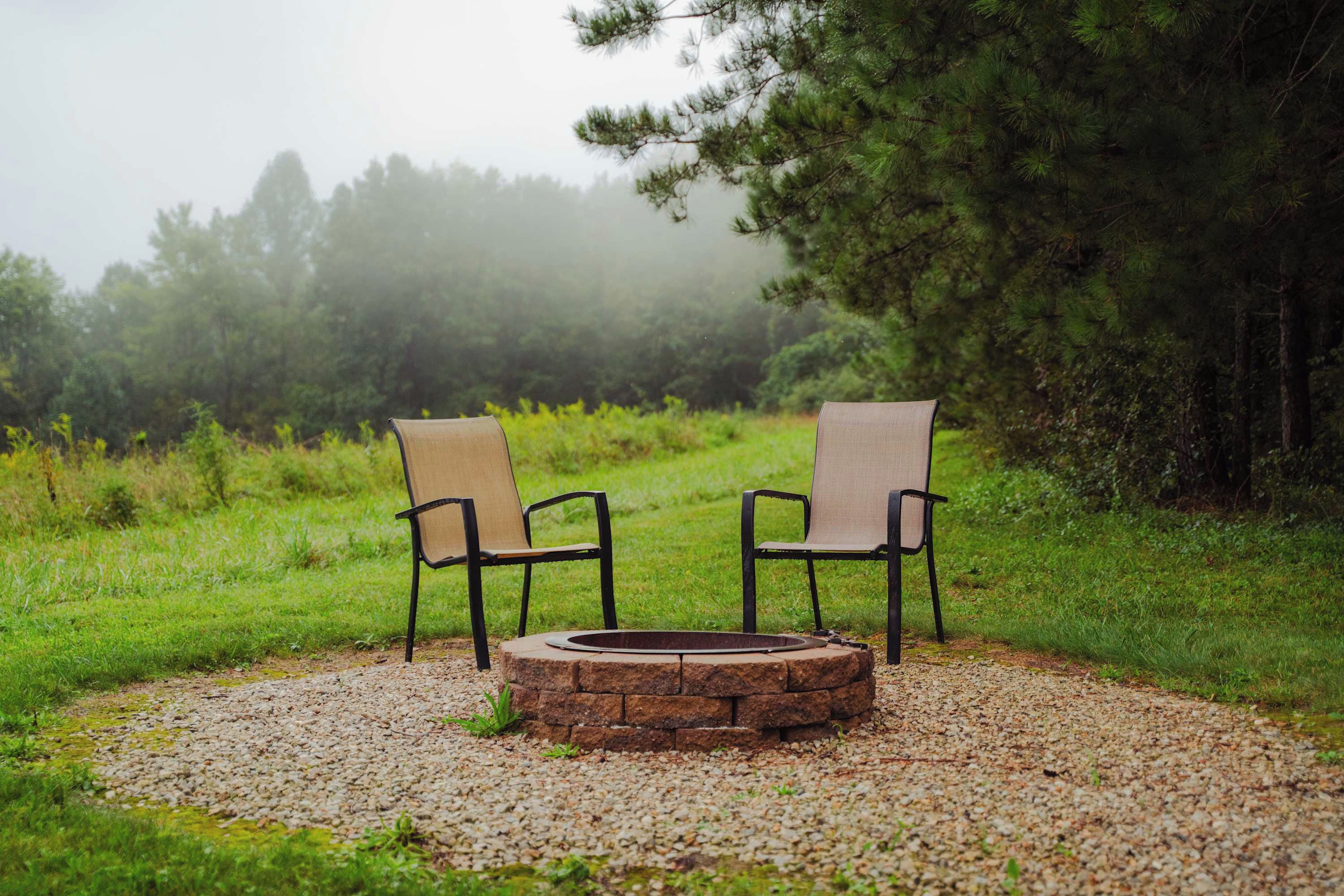 Two chairs facing a stone fire pit surrounded by gravel, set in a grassy area with trees in the background.