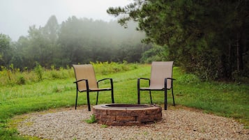 Two chairs facing a stone fire pit surrounded by gravel, set in a grassy area with trees in the background.