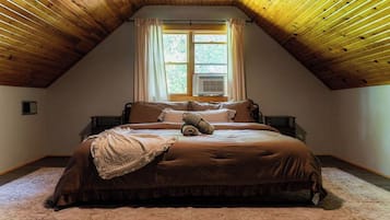 A bedroom with a sloped ceiling, a bed with a brown bedspread, a window with curtains, and an air conditioning unit.