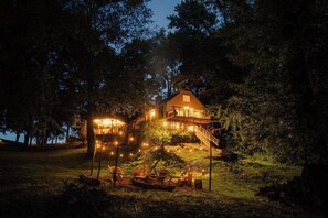 A house with a balcony, surrounded by trees, and illuminated by string lights. - Waterfront Lake Views Villa With Hot tub in Hocking Hills (Logan)