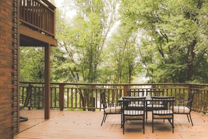 Um deck de madeira com mesa e cadeiras, rodeado por um corrimão de madeira, com vista para uma área arborizada.