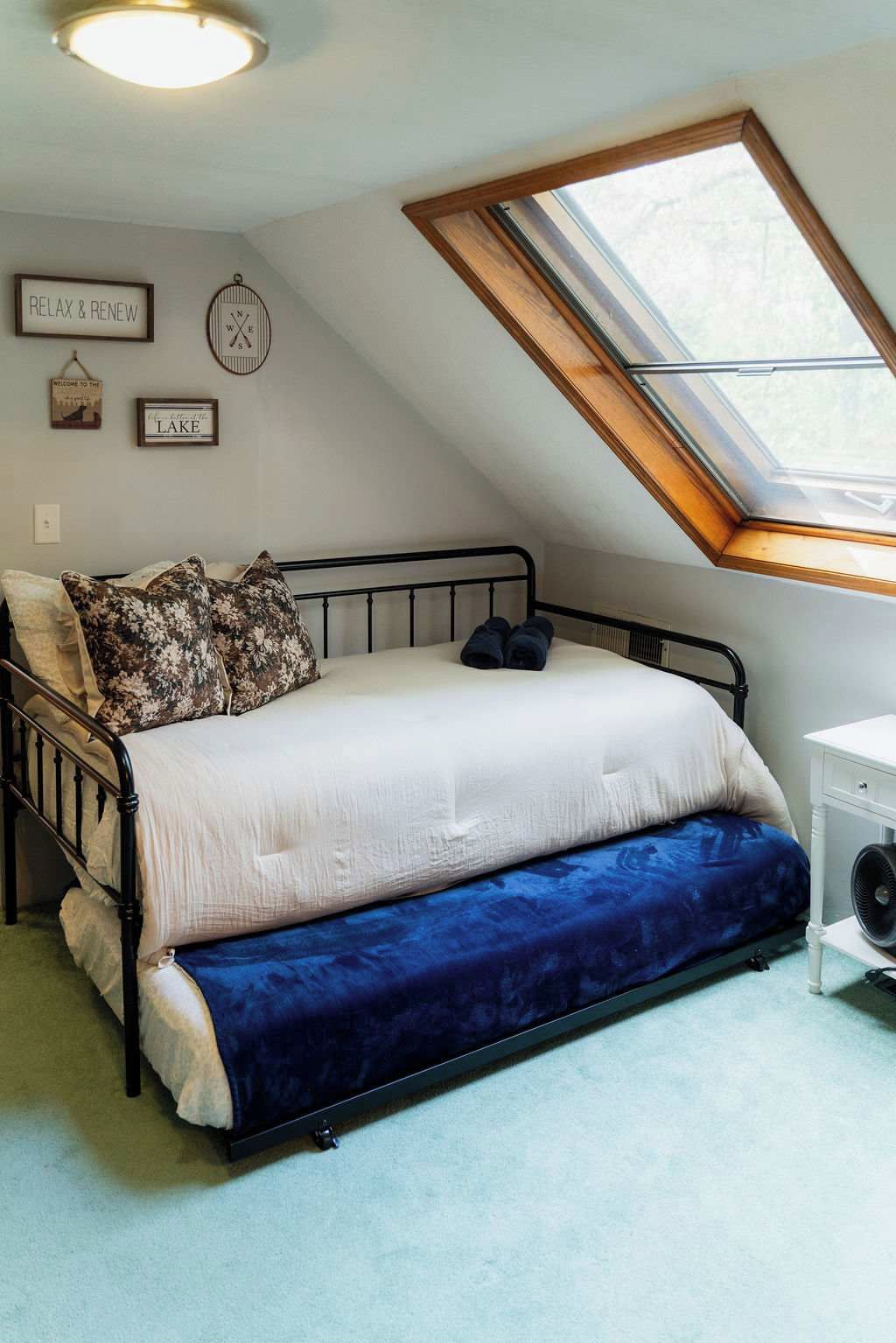 A bedroom with a sloped ceiling, a skylight, a bed with a white and blue duvet, and a small white bedside table.