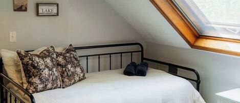 A bedroom with a sloped ceiling, a skylight, a bed with a white and blue duvet, and a small white bedside table.