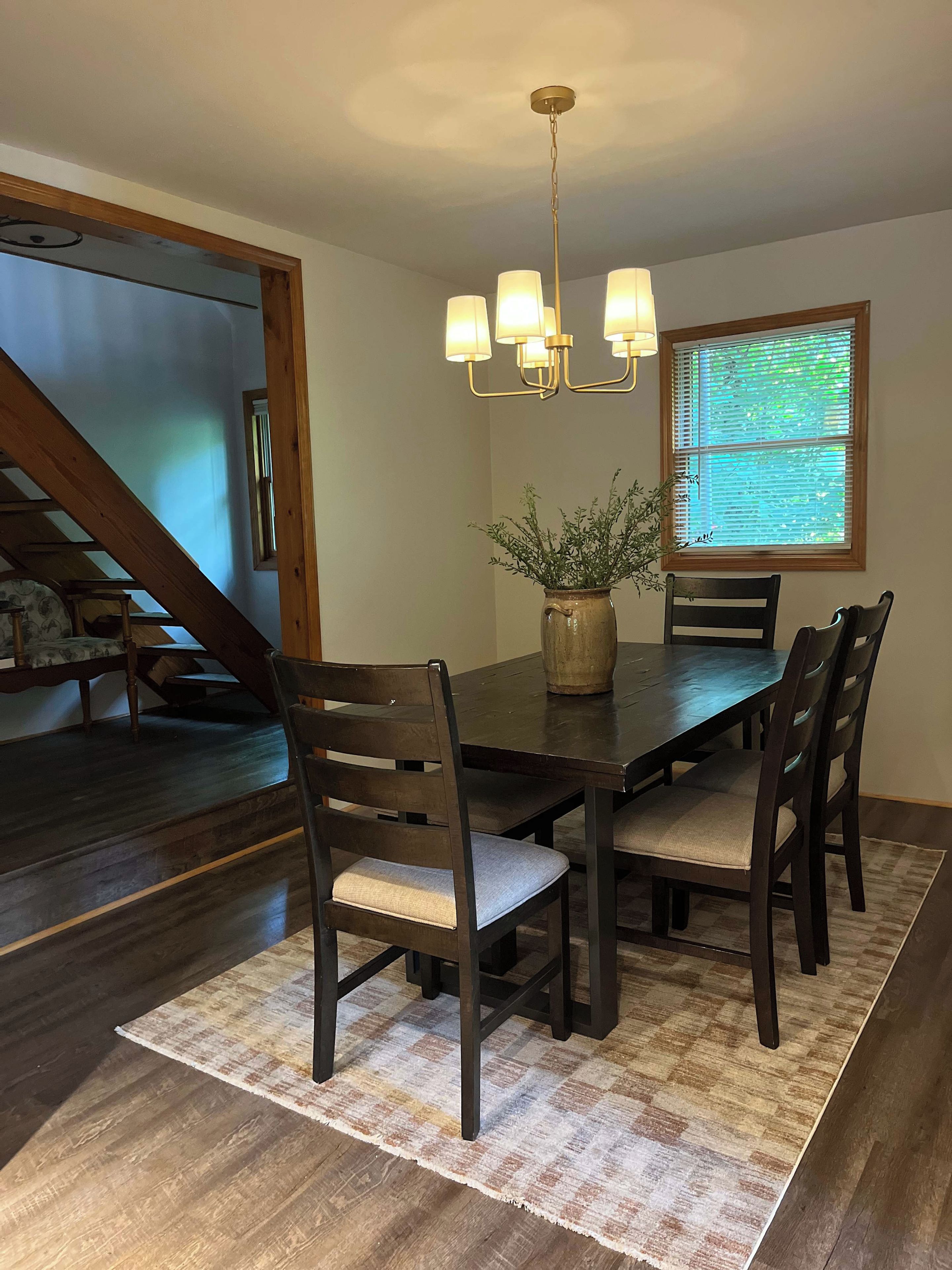 A dining area with a dark wooden table, four chairs, a chandelier, and a window with blinds.