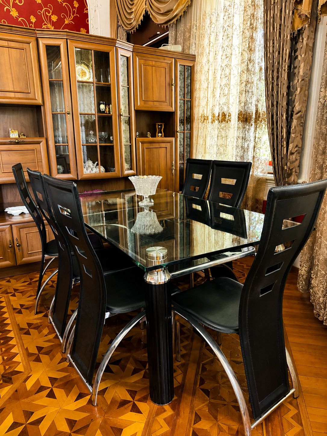 Un área de comedor con una mesa de vidrio rodeada de sillas negras, armarios de madera con puertas de vidrio y cortinas estampadas.