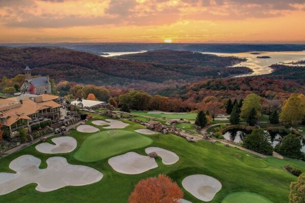 A golf course with bunkers, a clubhouse, and a scenic view of a lake and forest.