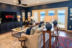 A living room with a sofa, coffee table, TV, and a view of the outdoors. - Sunset Shores: Waterfront Views + Pool, Branson & Golf Access (Hollister)