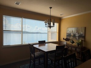 A dining room with a large table and chairs, a chandelier, a painting on the wall, and a view through the window.