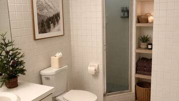 A bathroom with white tiled walls, a toilet, a sink, a framed picture, a shower with a glass door, and shelves with towels and toiletries.