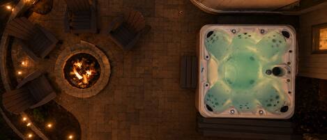 A hot tub with a fire pit, surrounded by Adirondack chairs, and string lights.