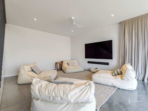 A modern living room with a flat-screen TV mounted on the wall, two bean bag chairs, a ceiling fan, and a large window with curtains.