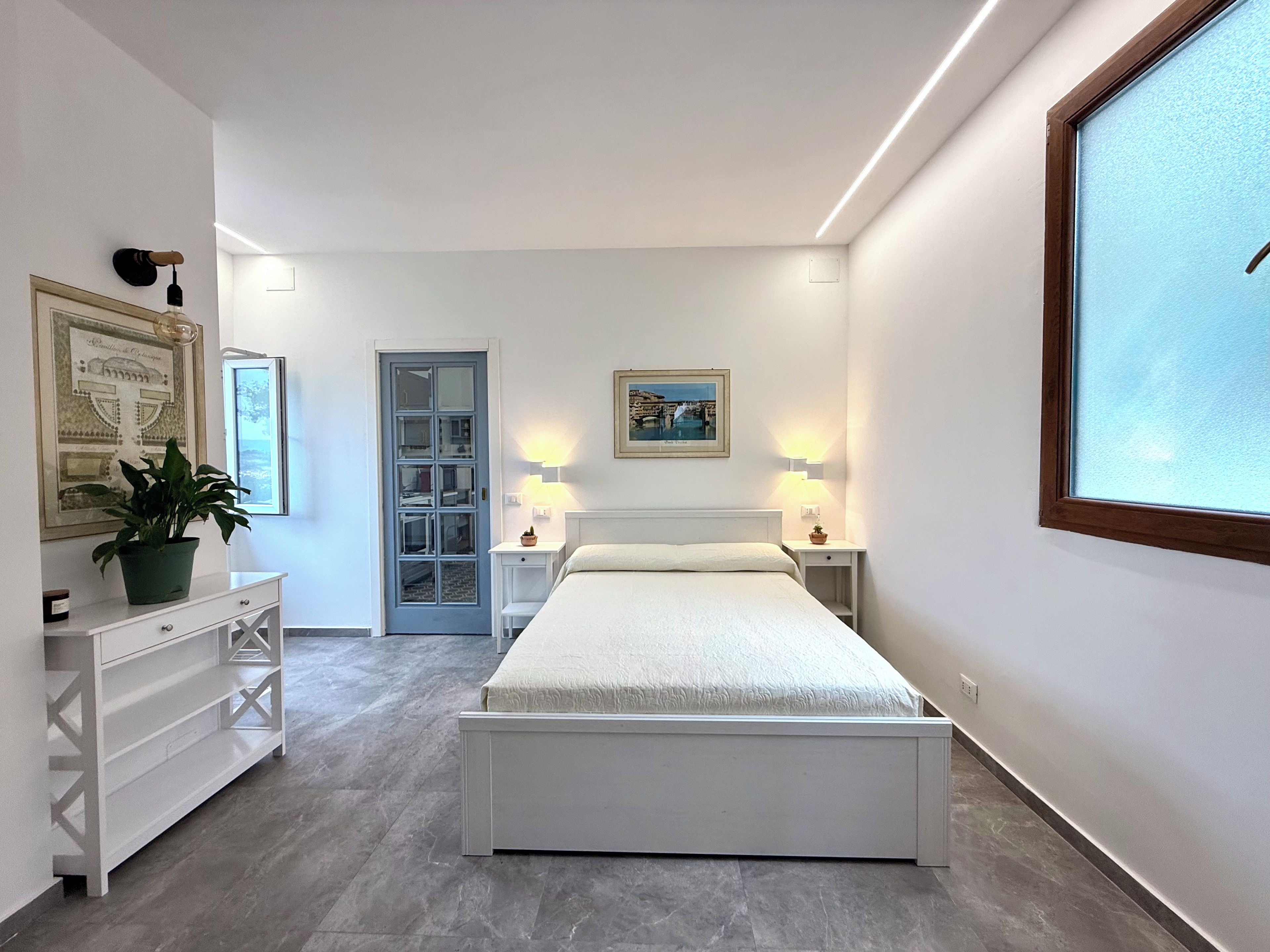 Une chambre à coucher moderne, avec un lit, des tables de chevet, une plante en pot et une porte donnant sur une autre pièce.