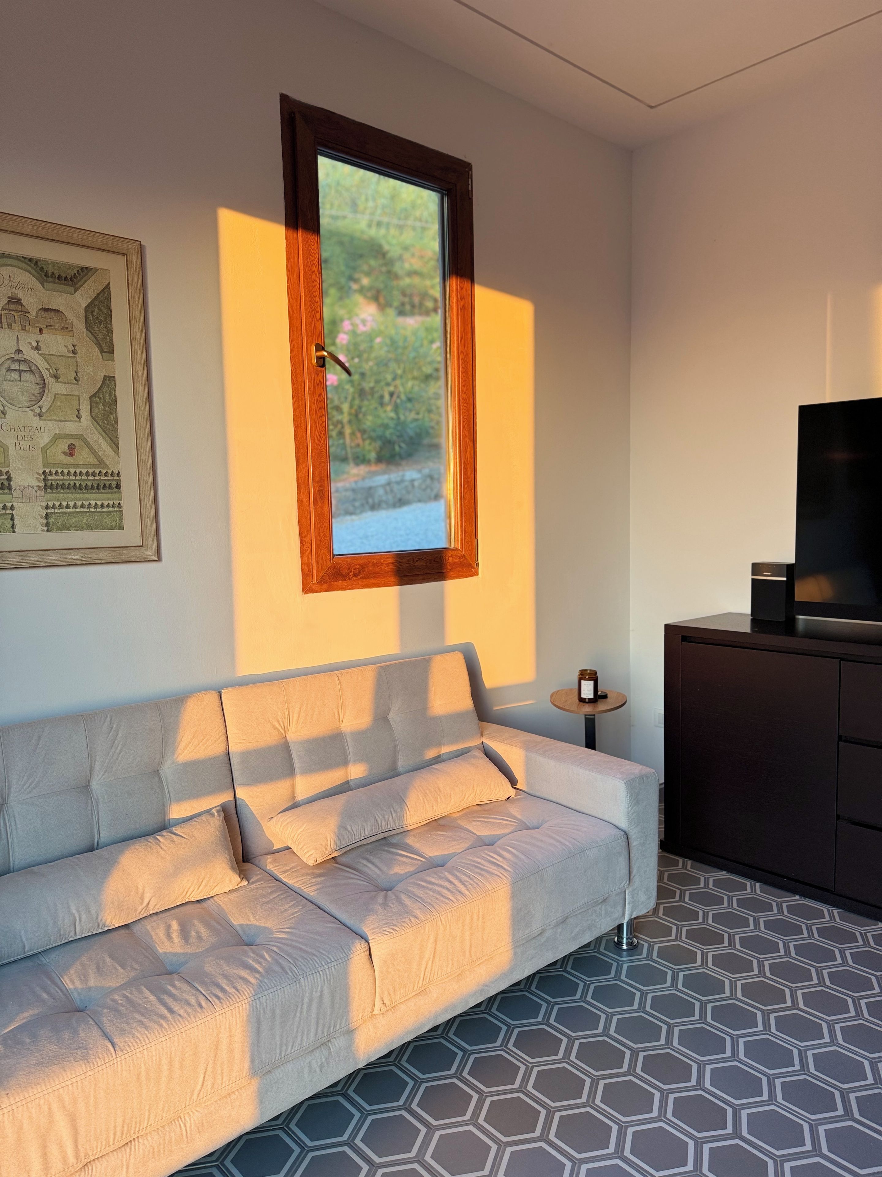 Ein modernes Wohnzimmer mit einer Couch, einem Fernseher auf einem Schrank, einem gerahmten Bild an der Wand und einem Fenster mit Blick auf Grünflächen.