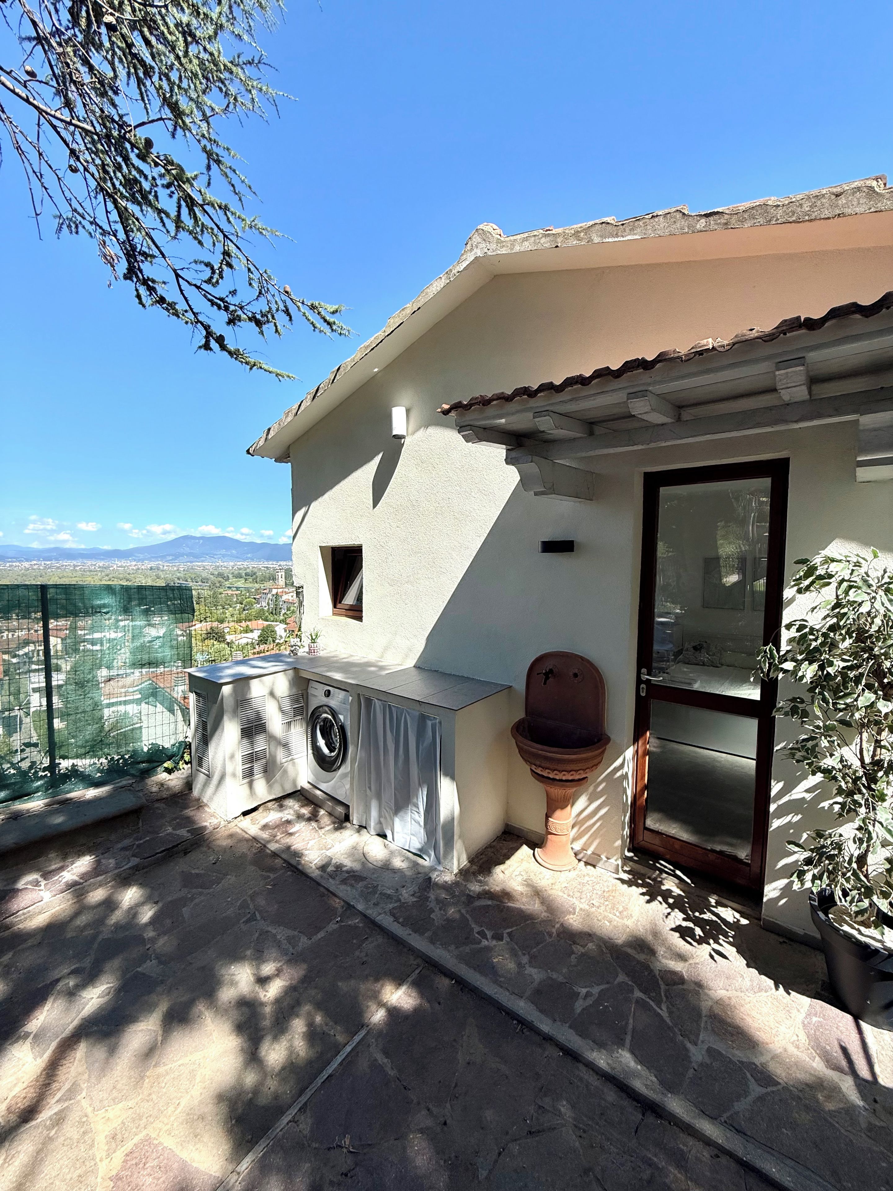 A balcony with a washing machine, a potted plant, and a view of distant mountains.