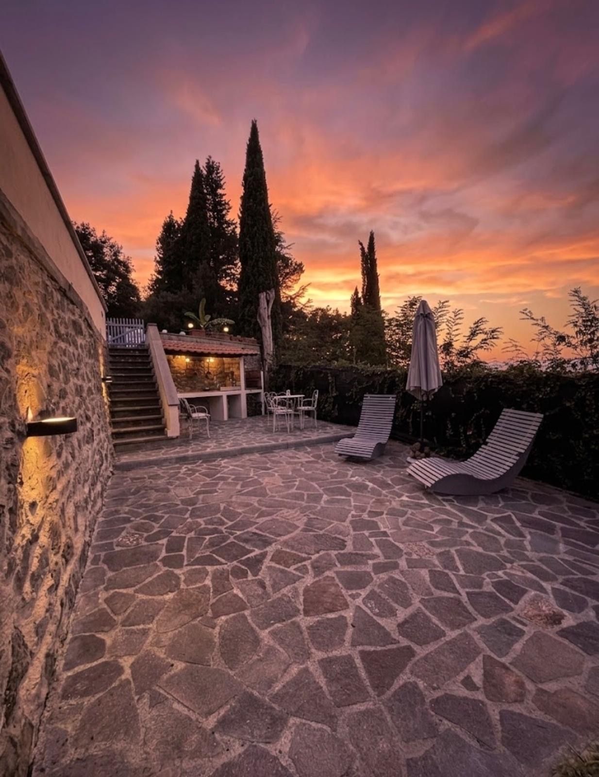 A stone patio with lounge chairs and a table, surrounded by trees and a building with a staircase.