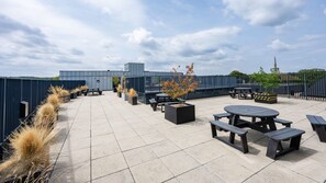 Eine Terrasse im Freien mit mehreren Tischen und Bänken, umgeben von einem Metallzaun, unter einem teils bewölkten Himmel.