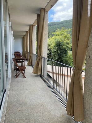 En balkon med et bord, stole, gardiner og udsigt til grøn vegetation.