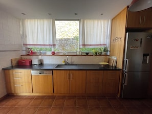A compact kitchen with wooden cabinets, a stainless steel refrigerator, and a window with curtains. - Countryside Sanctuary | Casa de Santiago Ponte de Lima (Facha)