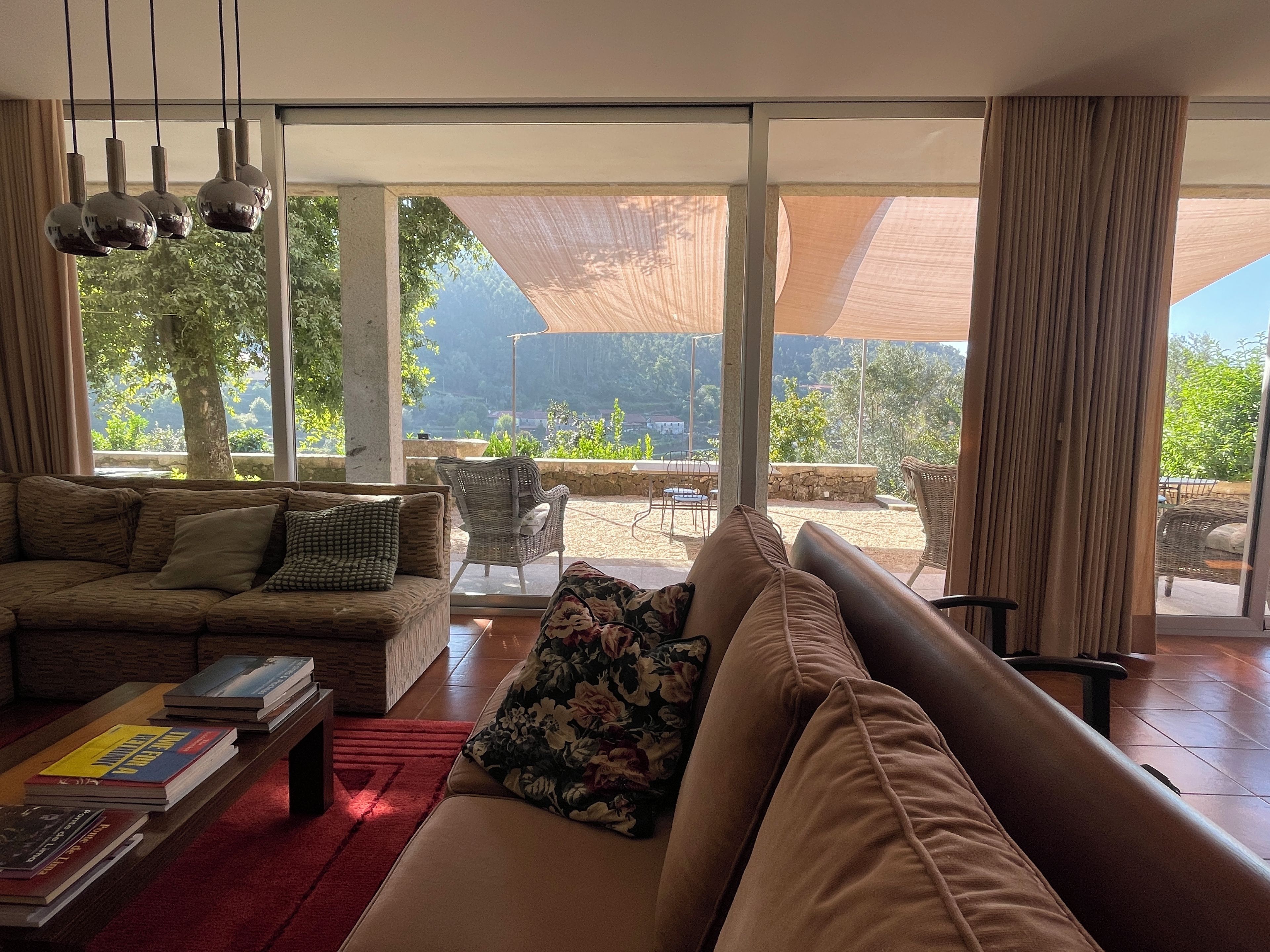 A spacious living room with a large sofa, a coffee table with books, and a view of the outdoors through large windows.