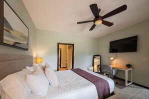 A hotel room with a bed, a desk, a chair, a ceiling fan, a TV, and a painting on the wall.