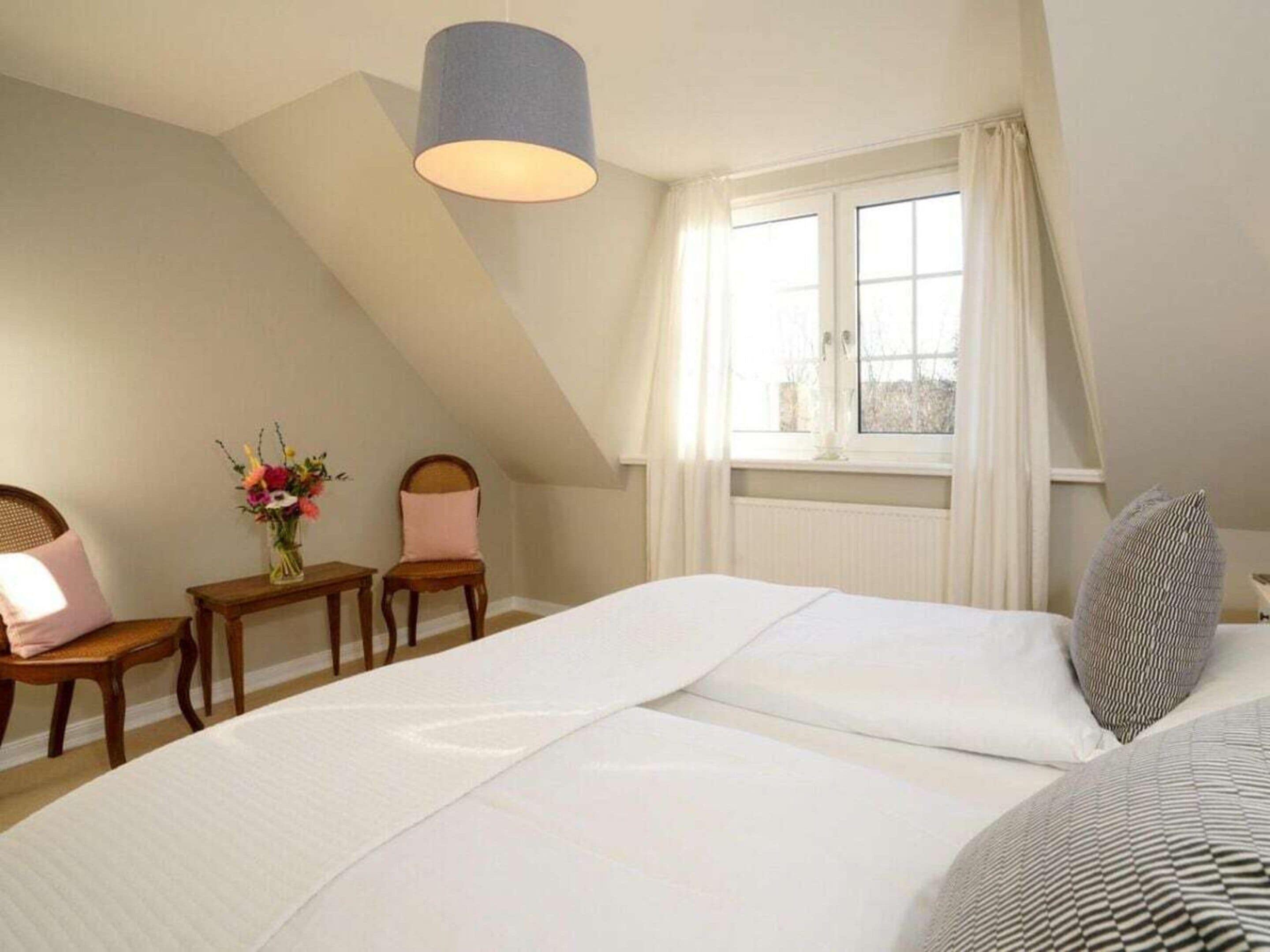 A bedroom with a sloped ceiling, a bed with white bedding, a bedside table with a vase of flowers, and a window with curtains.