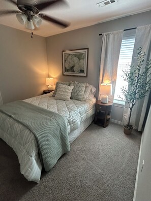 A bedroom with a bed, two bedside lamps, a ceiling fan, a window with curtains, and a potted plant.