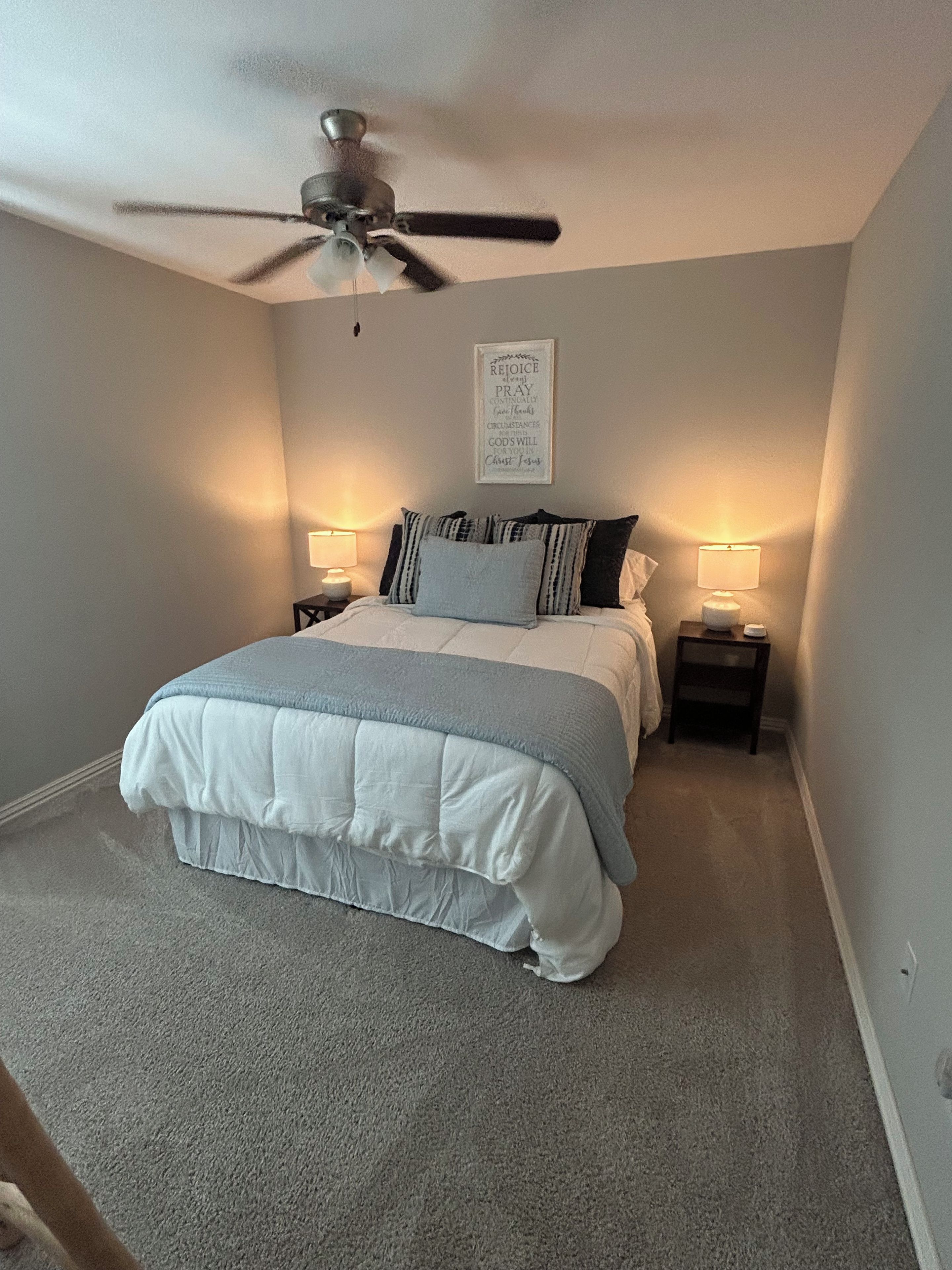 A bedroom with a bed, two bedside tables with lamps, a ceiling fan, and a motivational quote on the wall.