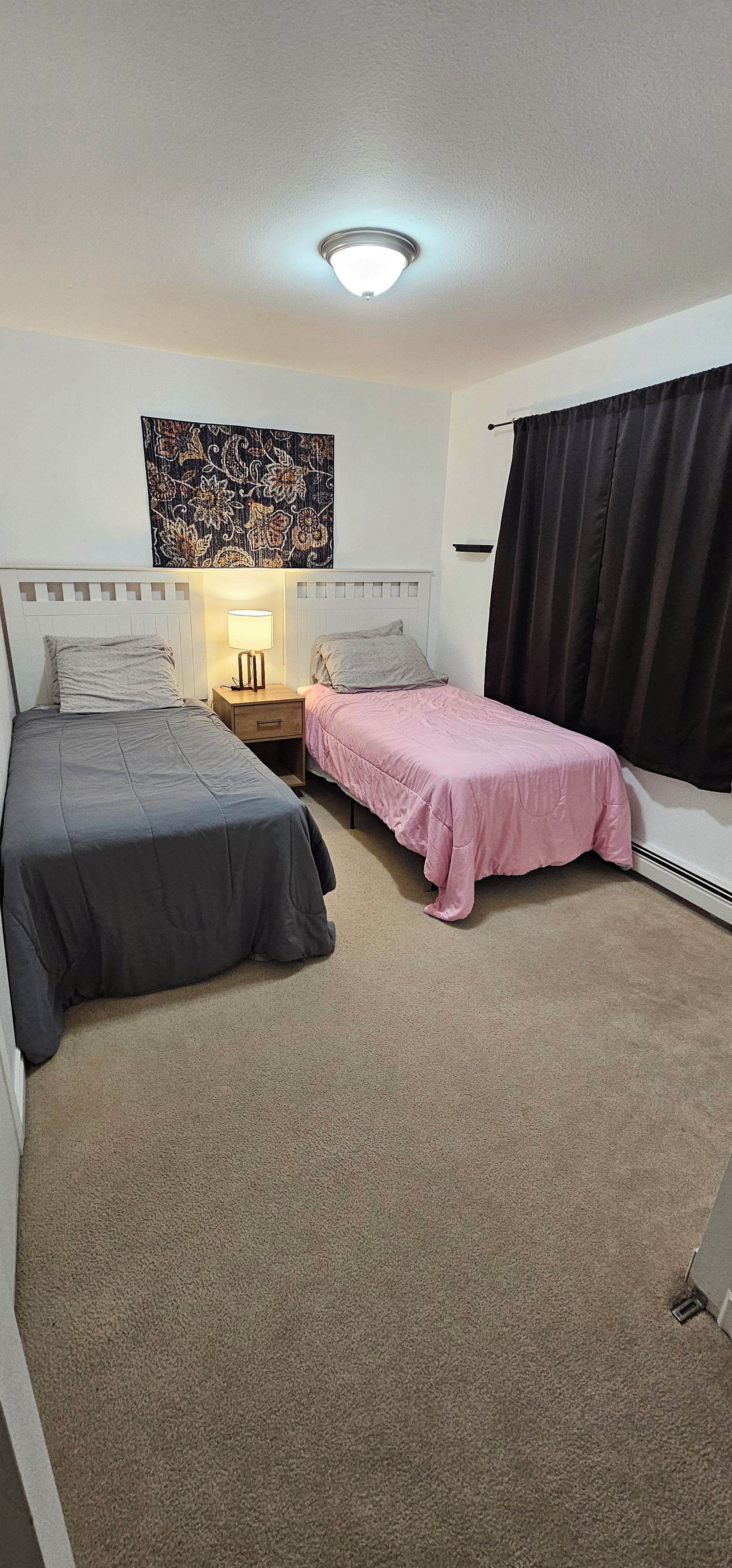 Een kamer met twee bedden, een nachtkastje met een lamp, een wanddecoratie en een raam met gordijnen.