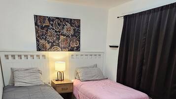 Een kamer met twee bedden, een nachtkastje met een lamp, een wanddecoratie en een raam met gordijnen.
