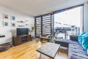 Ein modernes Wohnzimmer mit Holzboden, einer Couch, einem Couchtisch, einem Fernseher auf einem Holzschrank und einem großen Fenster mit Blick auf ein Gebäude.