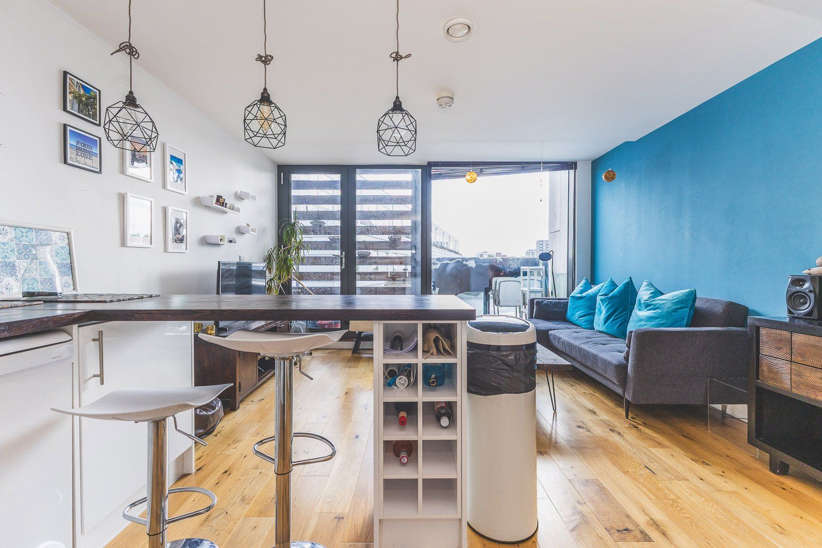 A modern apartment with a kitchen, dining area, and living room. There are hanging lights, a large window, and a blue accent wall.