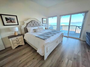 Ein Schlafzimmer mit einem großen Bett, einem Nachttisch, einer Kommode und Blick auf den Ozean durch eine Glastür.