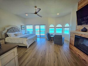 Quarto com cama, ventilador de teto, lareira e vista para o oceano através de grandes janelas.