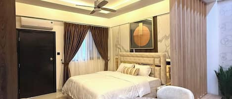 Un dormitorio moderno con cama, mesitas de noche, una mesa redonda con sillas, un ventilador de techo y un ventanal con cortinas.